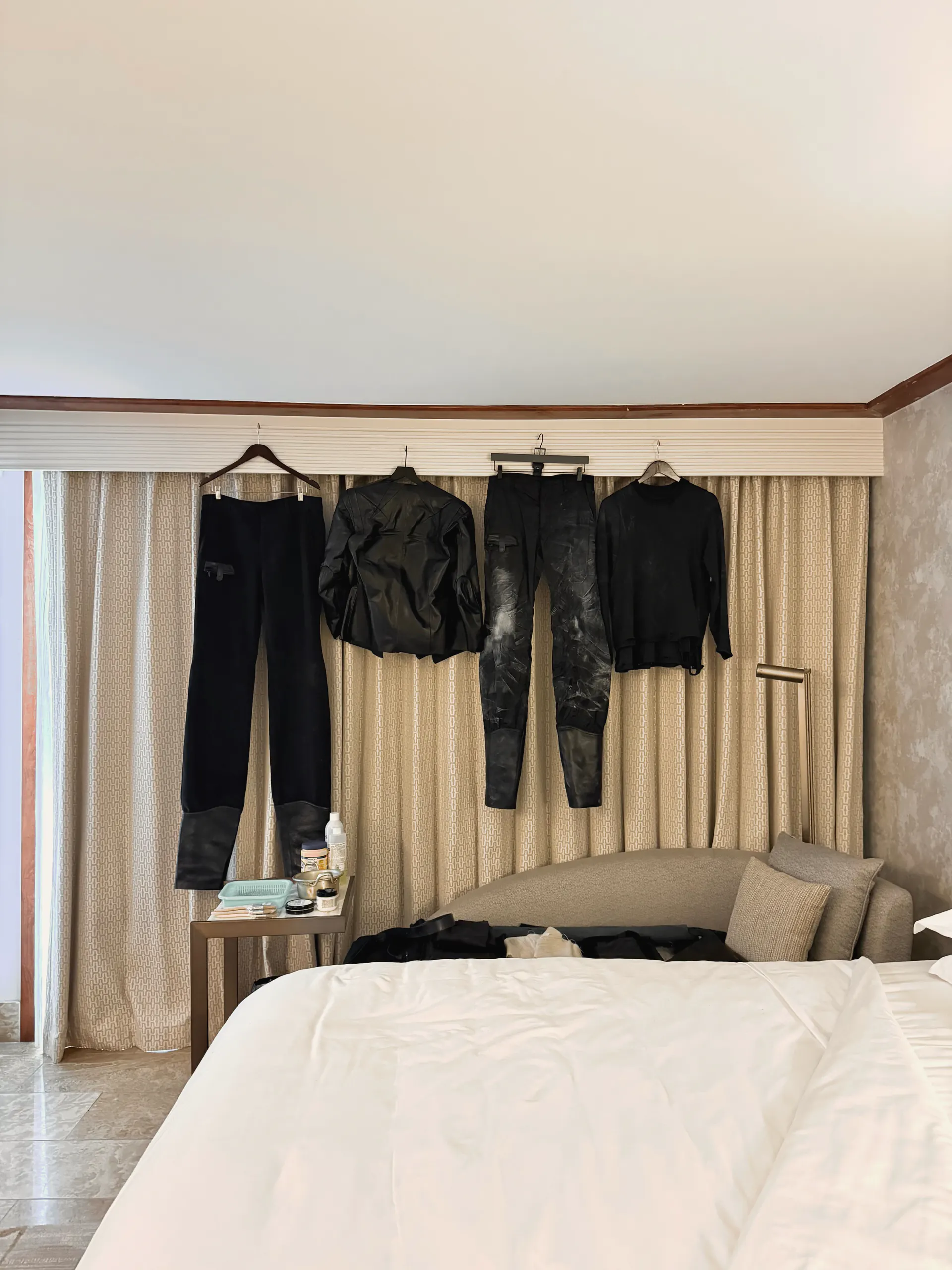 Four black garments hung on a curtain rail above a bed: trousers, a jacket, distressed leather trousers and a crewneck knit