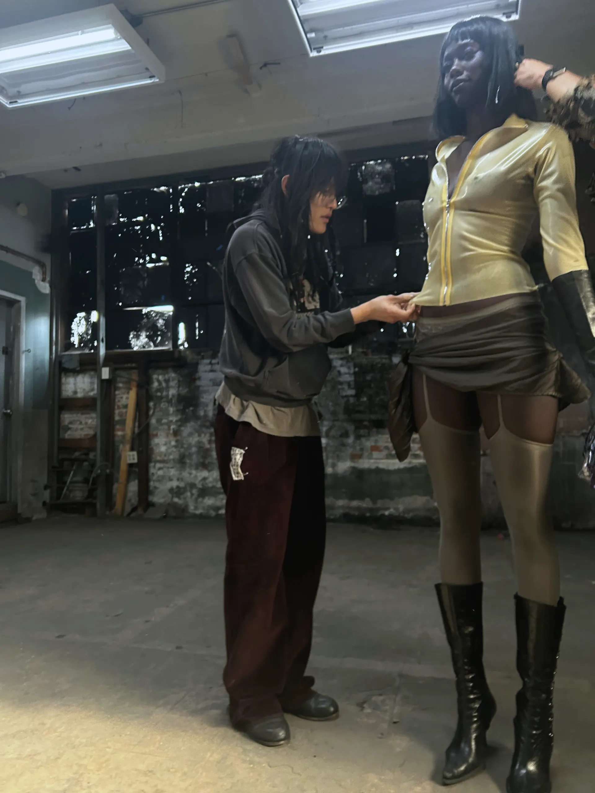 A fitting in a raw industrial space: one person adjusts the waist of a draped mini skirt on a model wearing a gold zip-front top, sheer hosiery, long black gloves and black knee boots.