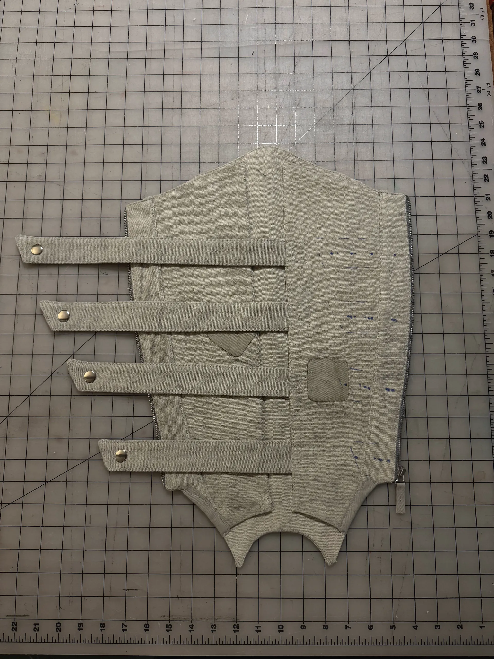 A pale canvas vest panel laid on a gridded cutting mat, with four strap extensions running off the left edge, each finished with a metal snap. A separating zipper runs down the right side, a small reinforcement patch is applied near the centre, and blue tailor's chalk marks dot the upper chest.