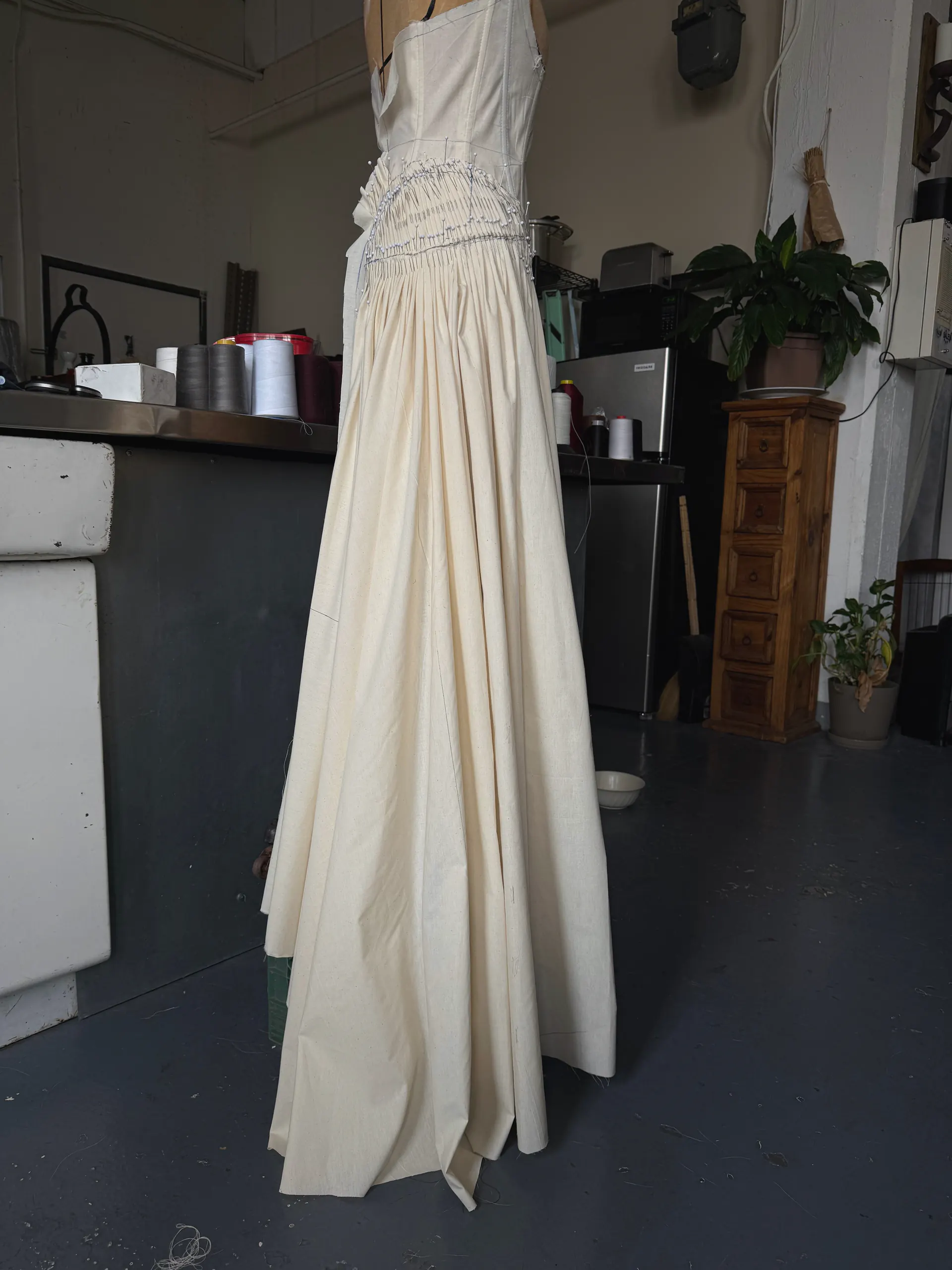 Cream cotton toile gown on a dress form, side view, gathered hip panel pinned in progress, in the studio.