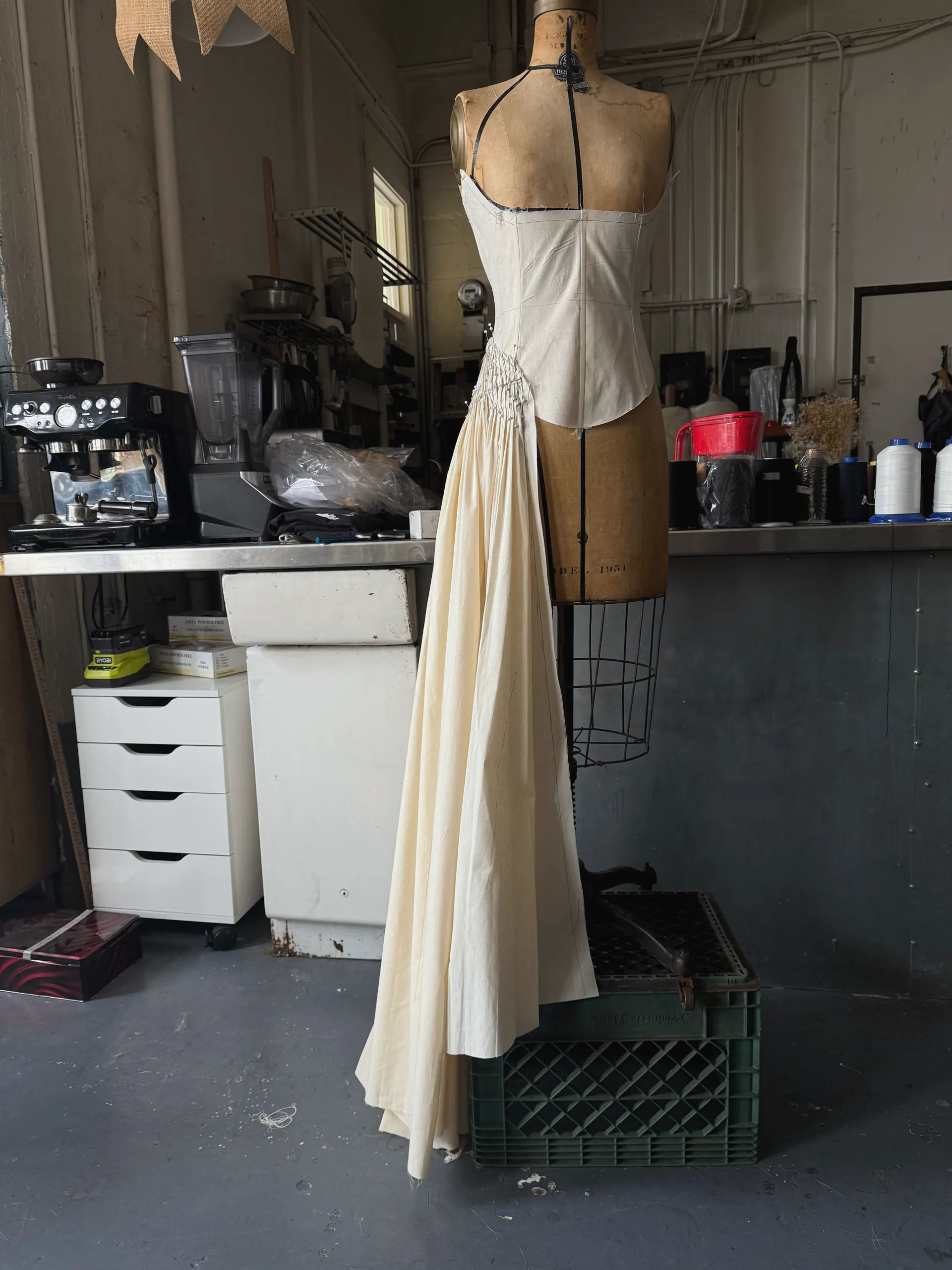 Cream cotton toile gown on a wire dress form in the studio, corseted bodice with one draped skirt panel falling to the floor.