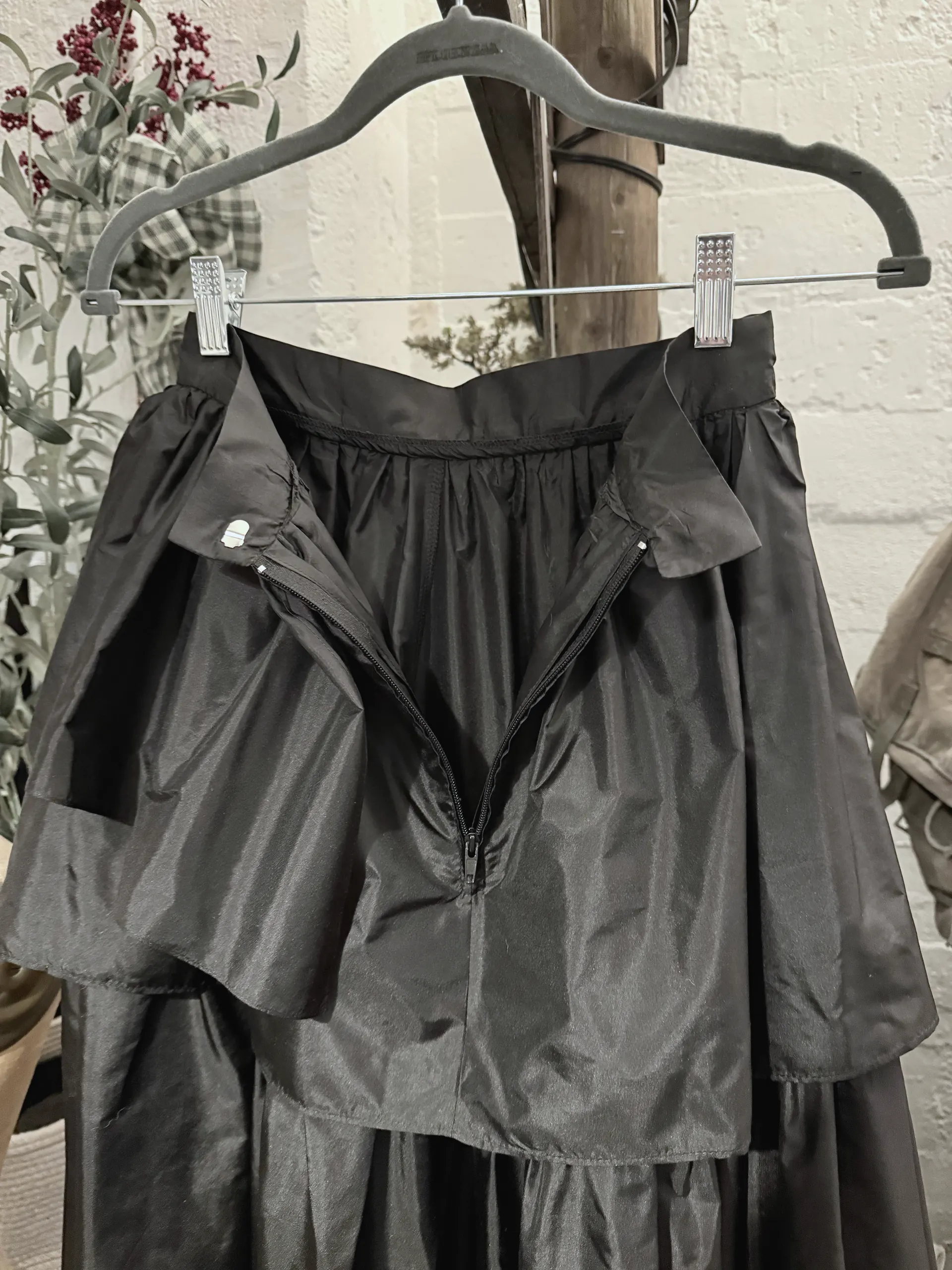 Close view of the waistband, open zip and gathered tiers of a black taffeta skirt on a hanger.