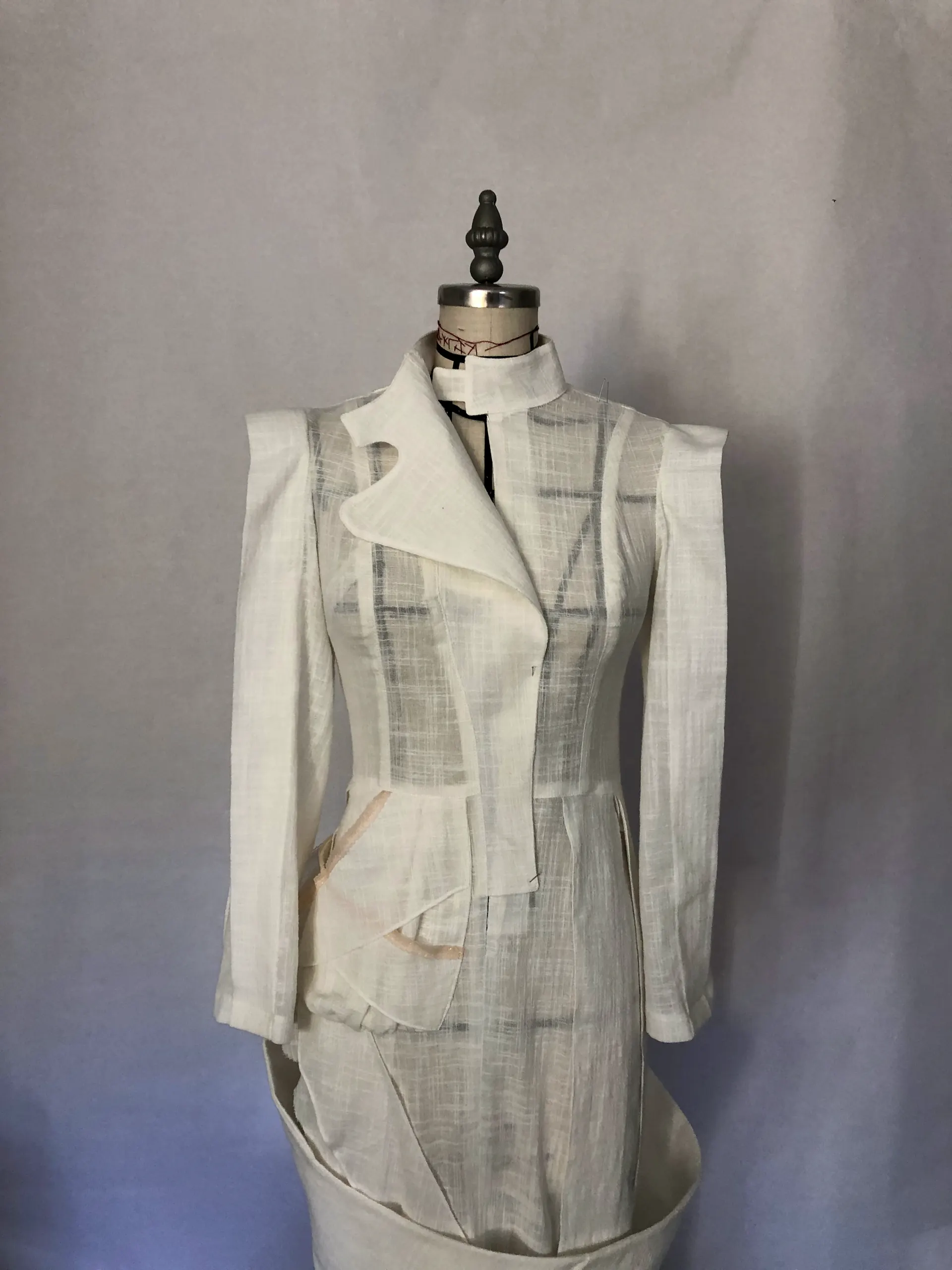 Sheer white tailored jacket with structured shoulders, stand collar and a peplum, with a matching skirt, on a dress form against a grey backdrop.