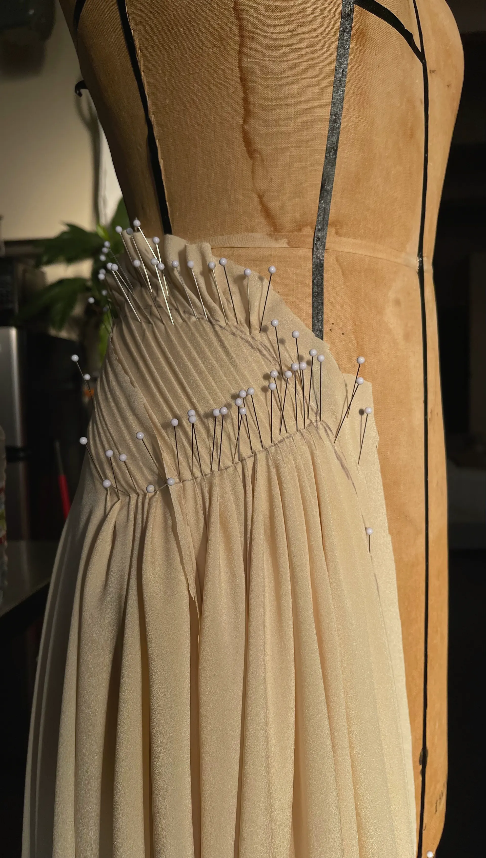 A cream sheer fabric is worked in progress on a linen dress form, the bodice gathered into dense fine pin-tucks along a curved yoke and held with rows of pins, the skirt falling away below in soft vertical folds.