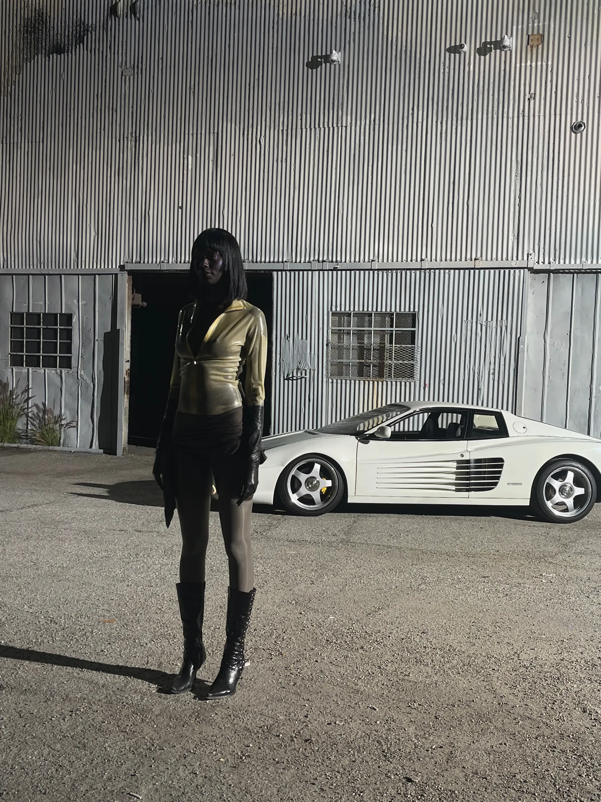 Model standing in a gravel lot in front of a corrugated metal building, wearing a gold satin zip-front top, a black skirt, sheer hosiery, long black gloves and laced black knee boots, with a white sports car parked alongside.