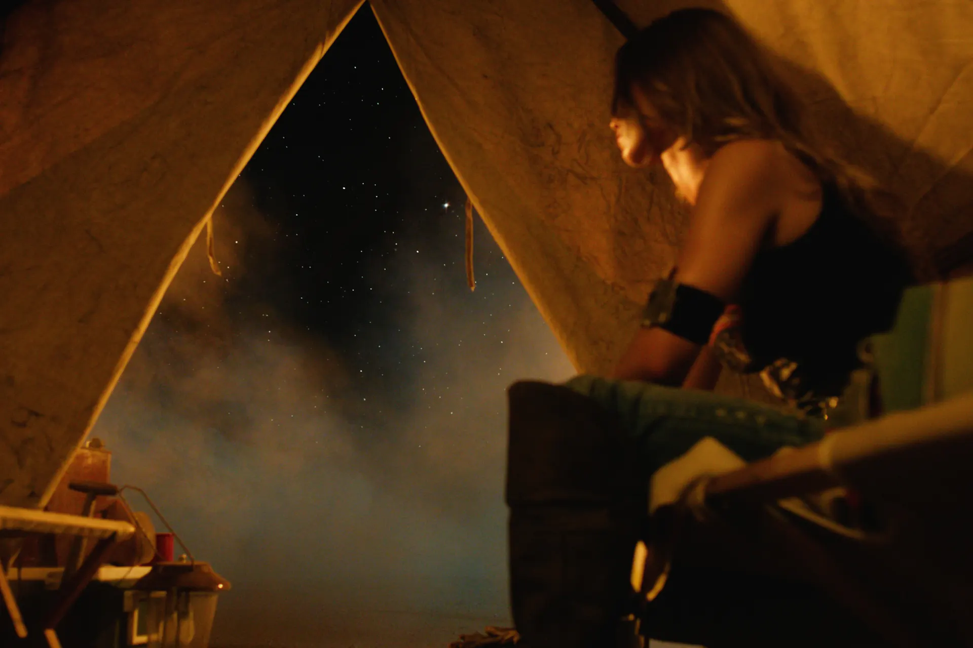 Inside a canvas tent lit warm at night, a figure in a black tank with a wide armband sits at the edge of the frame in profile, while the open tent flap reveals a star-filled sky and drifting haze beyond.
