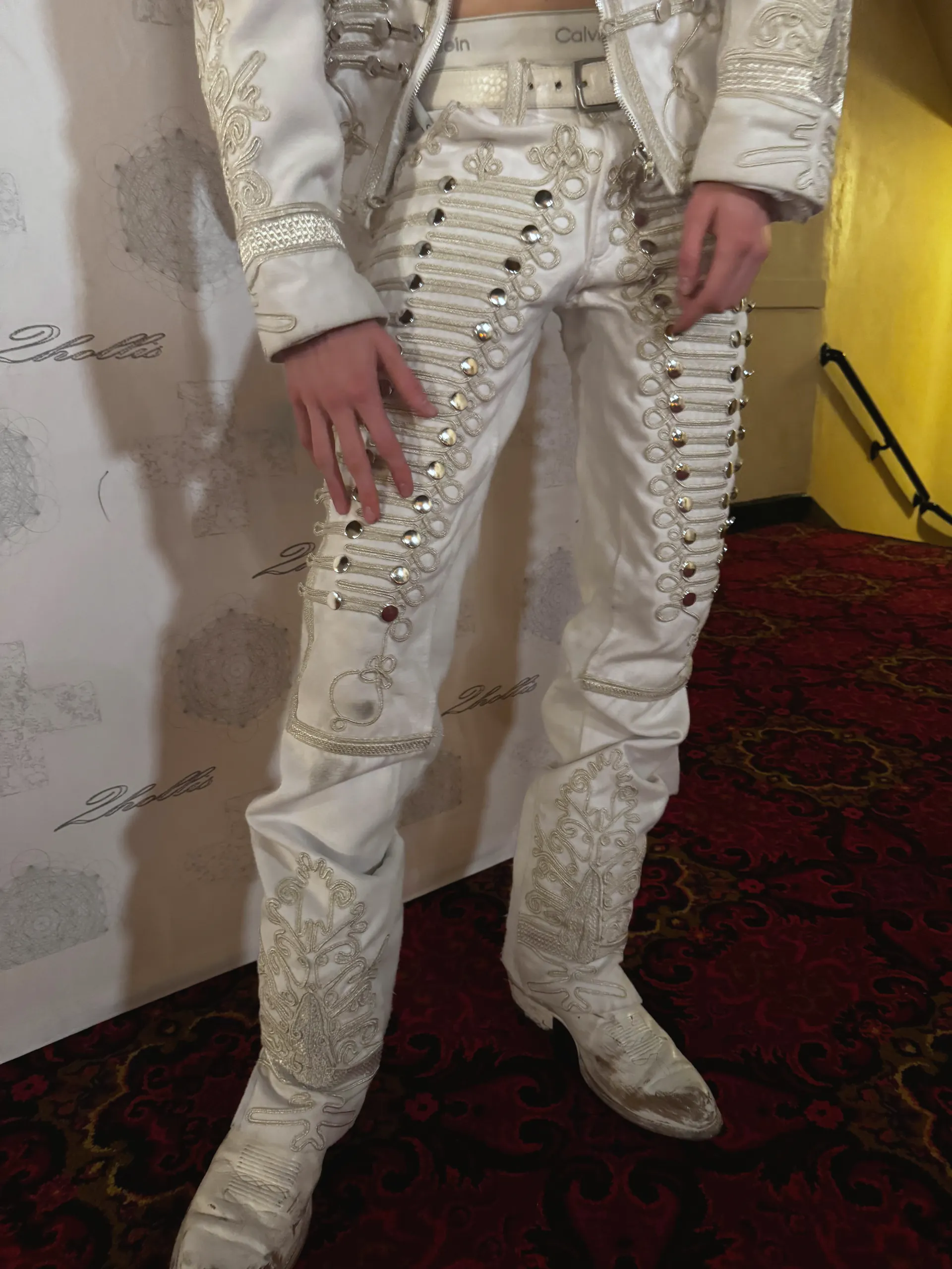 White jacket and trousers with gold cord embroidery, mirrored studs and matching boots, photographed from the chest down against an event backdrop.