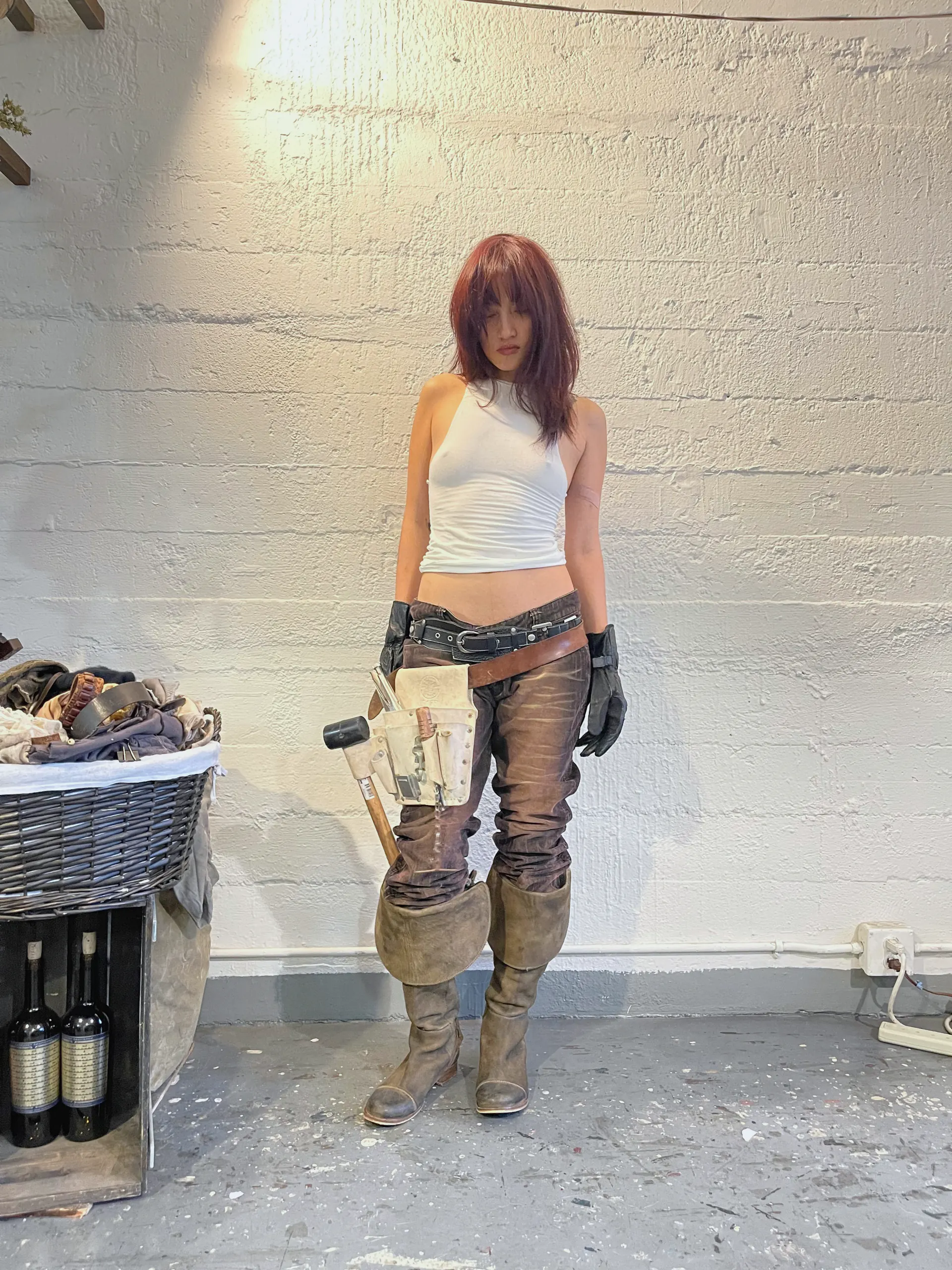 A model stands against a white painted brick wall in a white ribbed tank and heavily distressed brown trousers, styled with a doubled belt, a leather tool holster carrying a mallet, black gloves and tall worn boots. A basket of garments and a crate of bottles sit at the edge of the frame.