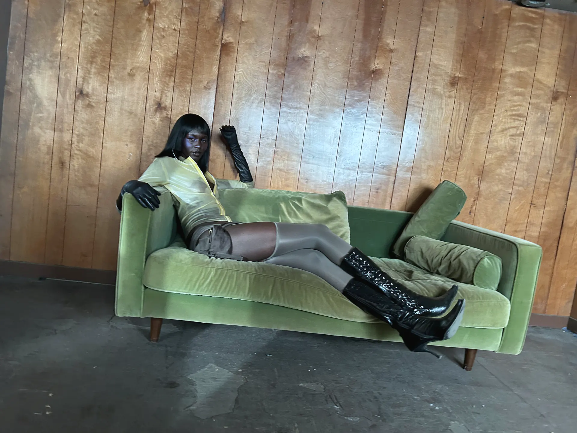 Model reclining full length on a green velvet sofa against a wood-panelled wall, wearing a gold satin top, a short draped skirt, sheer thigh-high hosiery, long black gloves and black patent knee boots.