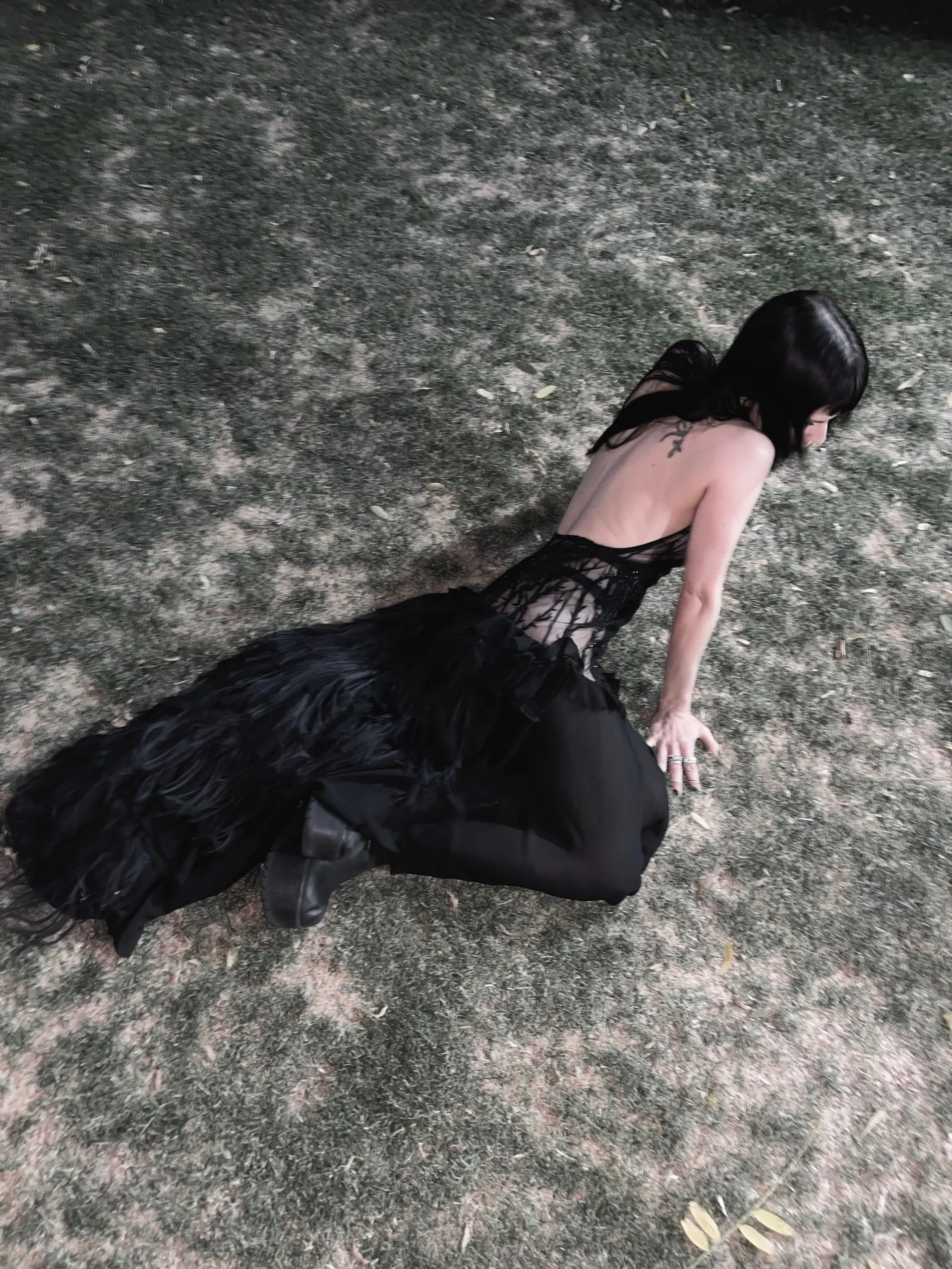 Person kneeling on grass at night in a black open-back gown, the lace-corset back panel and long ruffled train spread across the ground.