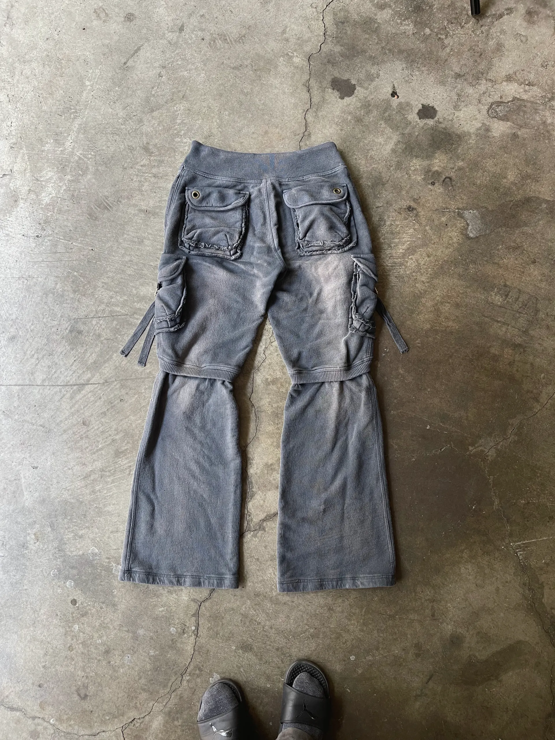 Back view of washed charcoal fleece cargo trousers with flap hip pockets, side cargo pockets and detachable lower legs, laid flat on concrete.