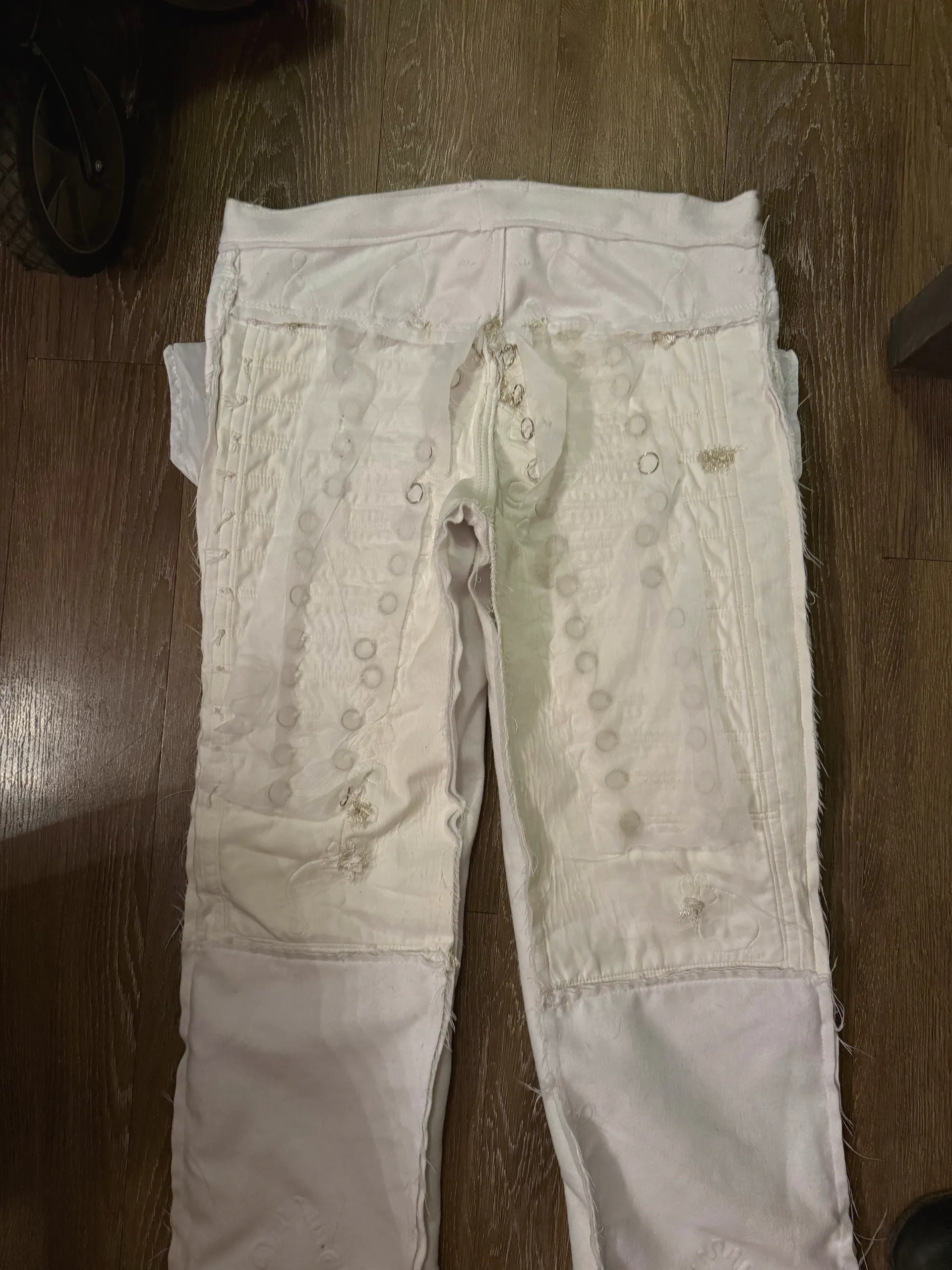 A pair of white trousers laid flat on a dark wood floor, worked over with dense repair: faint circular shadows run in columns down both legs, small hand-darned patches interrupt the surface, and a contrasting white panel is joined across each knee with a raw, fraying seam allowance left exposed down the outer leg.