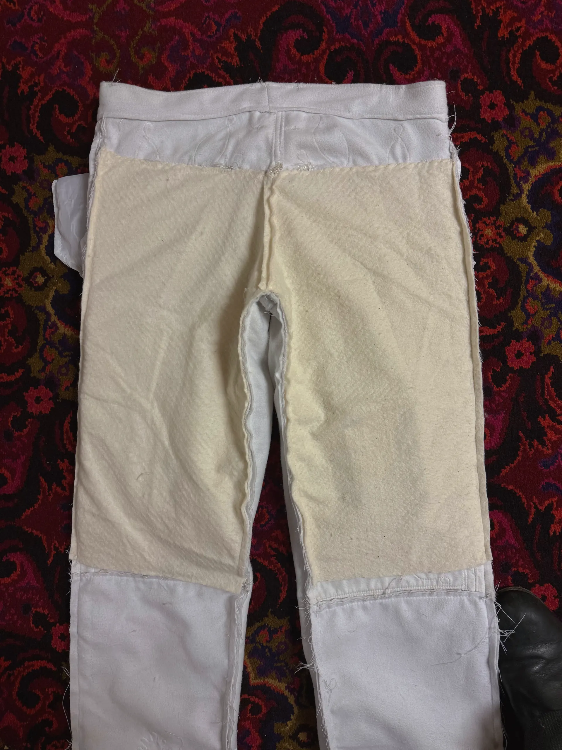 The same white trousers laid on a red patterned carpet with a cream, felted inner layer exposed across the seat and upper legs. The pale outer shell shows only at the waistband and the lower legs, and every joining seam is left raw with the allowance standing proud of the surface.