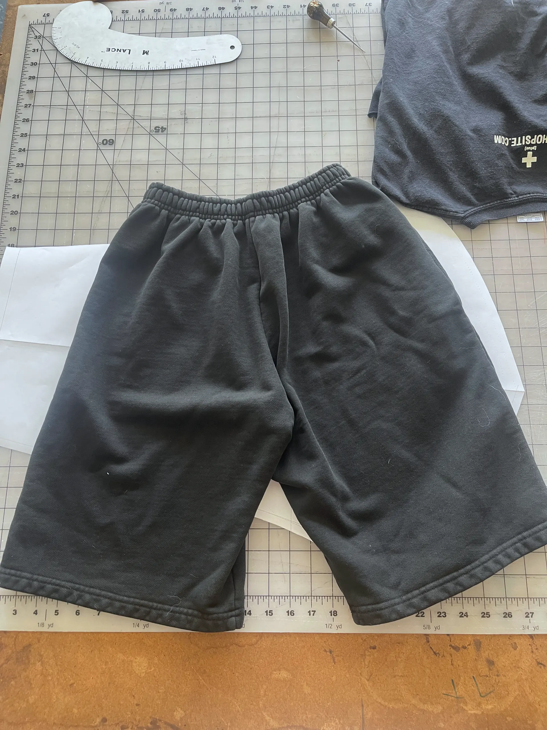 The same faded black fleece sweat shorts are shown flat from a second angle on a gridded cutting table, the ribbed elastic waistband gathered across the top and the wide legs spread to show their full width. Inch and yard markings printed on the mat run along the bottom of the frame.