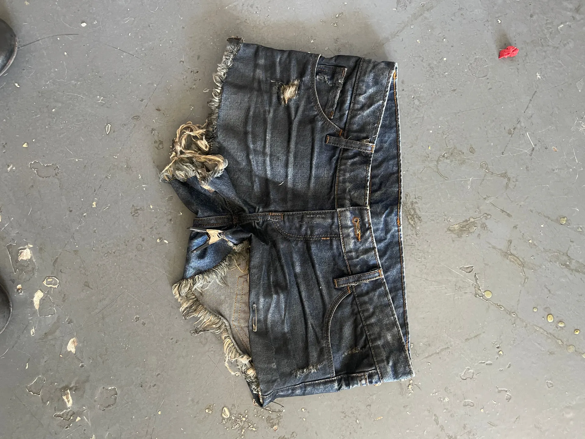 A pair of cut-off indigo denim shorts lies front side up on a scuffed concrete floor, the legs chopped short and left to fray into long pale threads. Heavy vertical fading runs down the front, the fly and belt loops are intact and the hems are unfinished.
