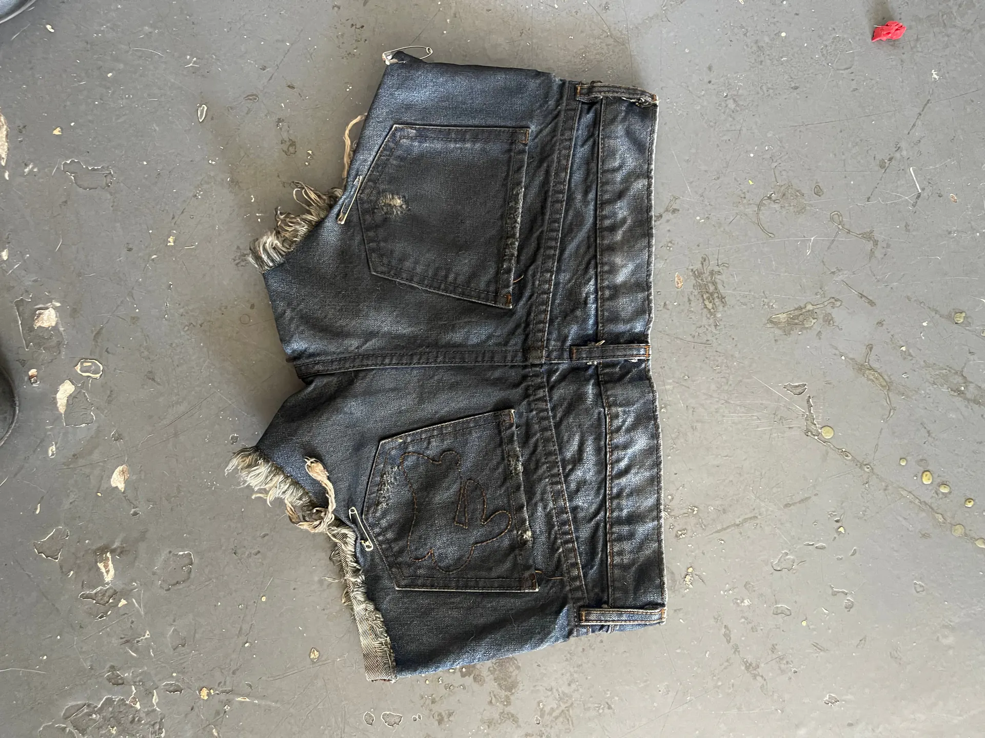 The same cut-off denim shorts are shown back side up on the concrete floor, with two patch back pockets, a felled centre seam and raw hems frayed into pale fringe. One pocket carries a curved double row of tonal topstitching and the denim is worn thin and dark across the seat.