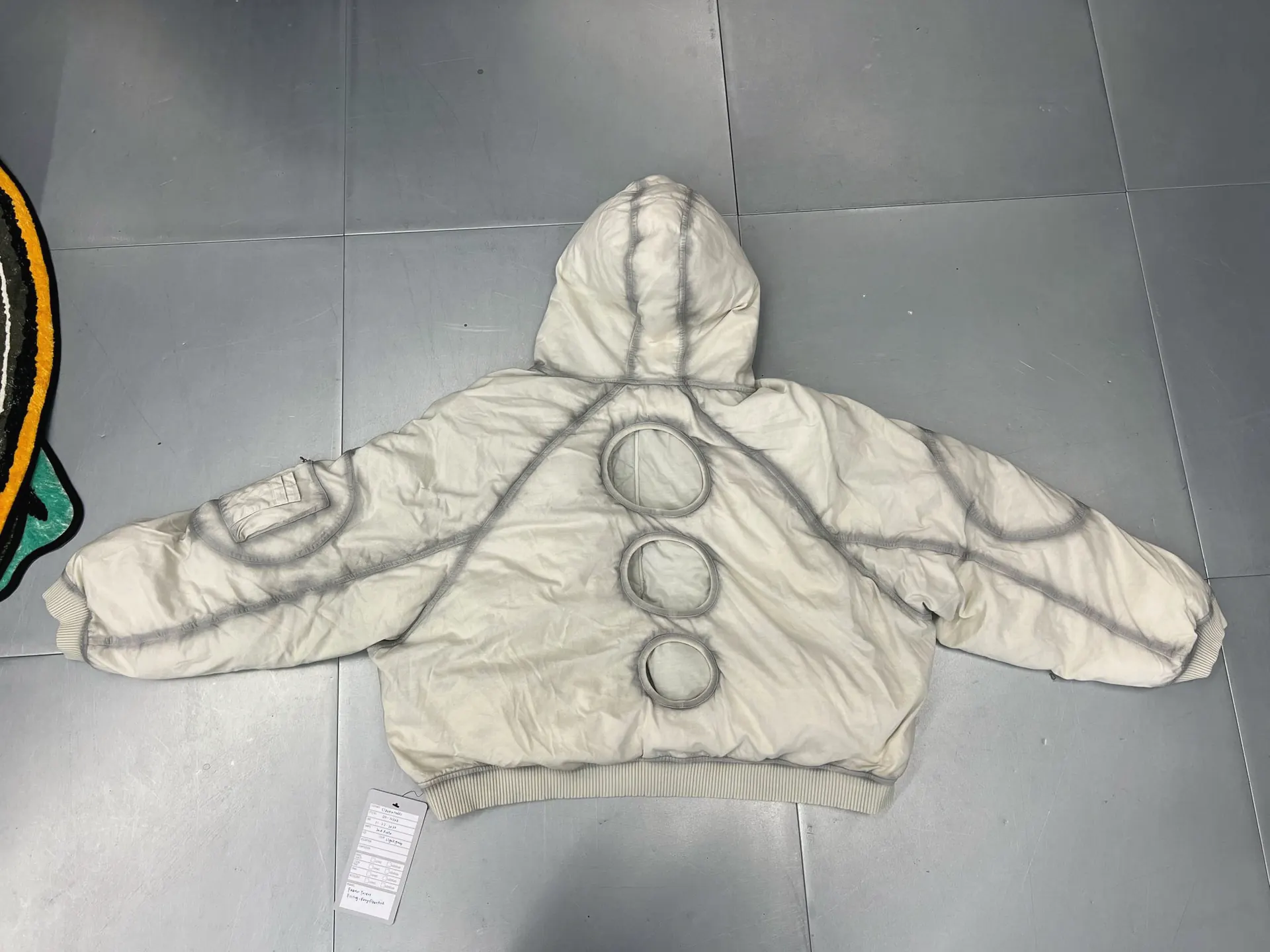 Cream padded hooded jacket, back view, with three circular openings down the centre back, contrast taped seams and ribbed cuffs and hem, laid on a tiled floor.