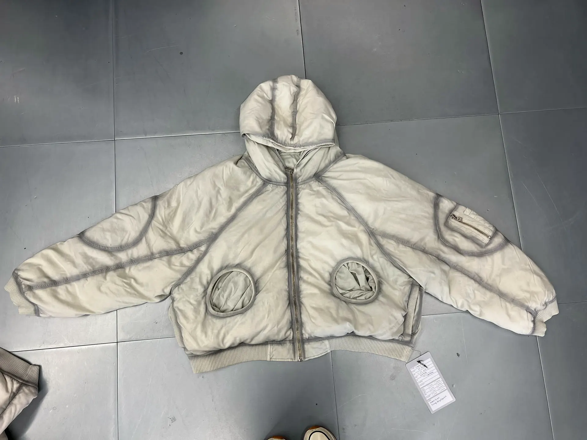 Cream padded hooded jacket, front view, zipped, with two circular openings at the front, a zip sleeve pocket, contrast taped seams and ribbed cuffs and hem, laid on a tiled floor.