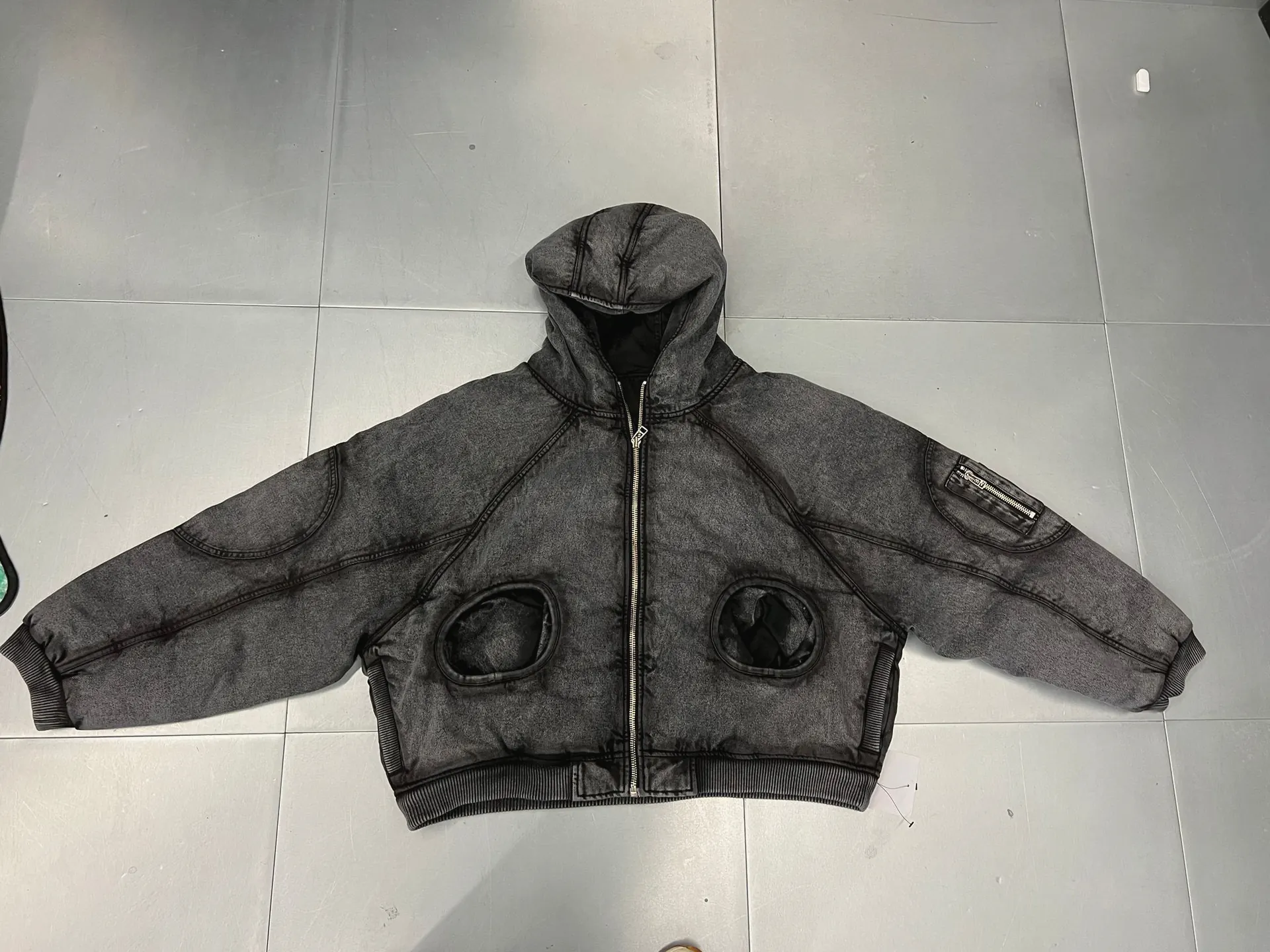 Washed black padded hooded jacket with curved seam panels, round set-in pockets, a sleeve zip pocket and ribbed cuffs and hem, laid flat on a tiled floor.