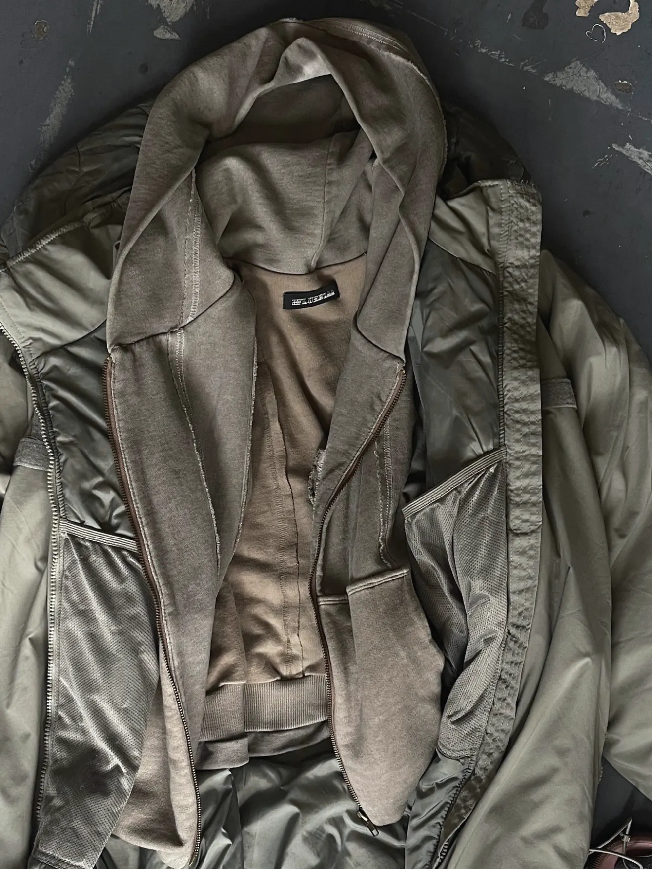 Washed brown hooded zip sweatshirt with raw-edged seams laid open over an olive nylon jacket with zip pockets, on a dark scuffed floor.