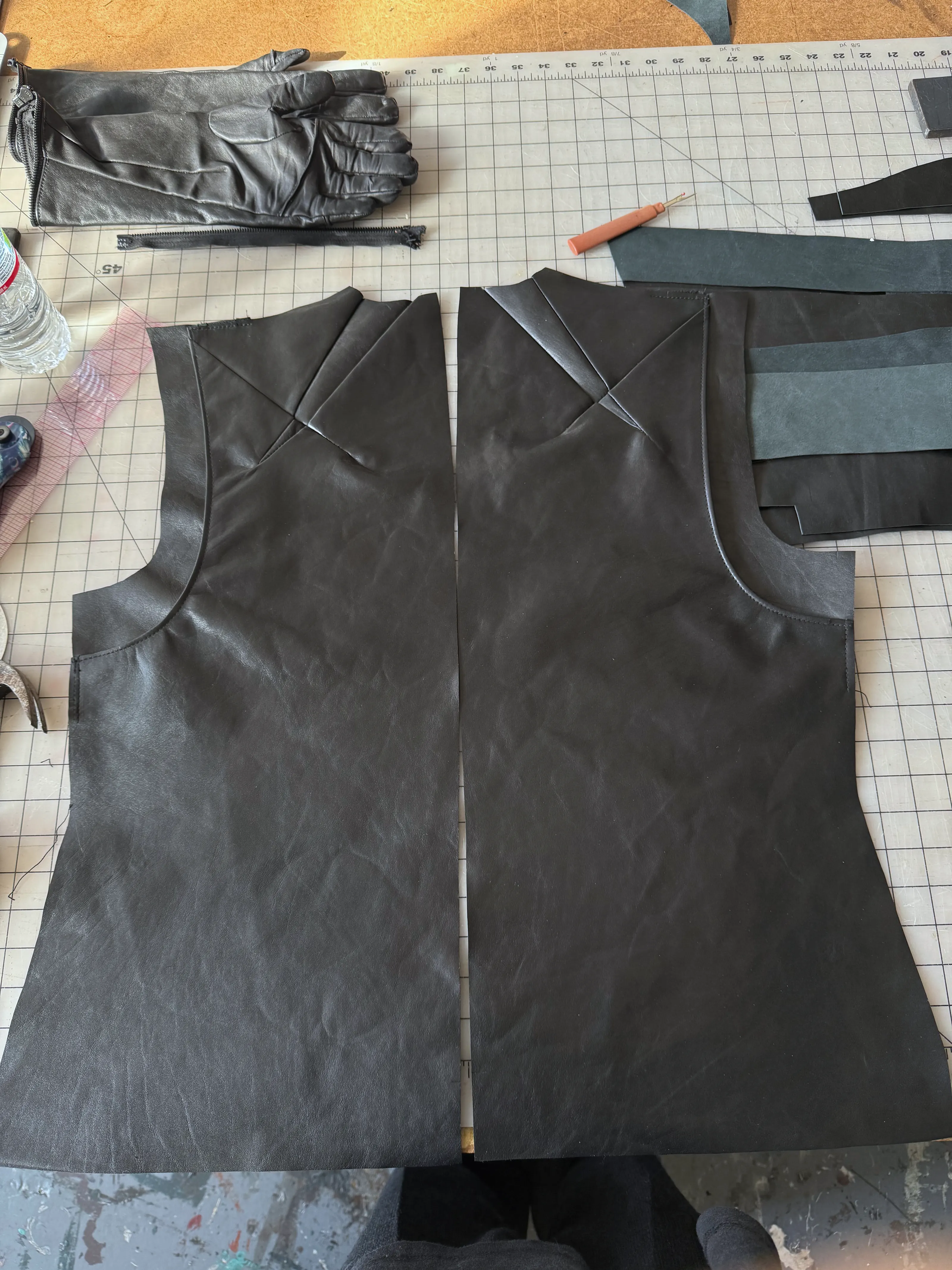 Cut black leather body panels with shoulder dart seams laid on a cutting table, a long leather glove and offcuts alongside.