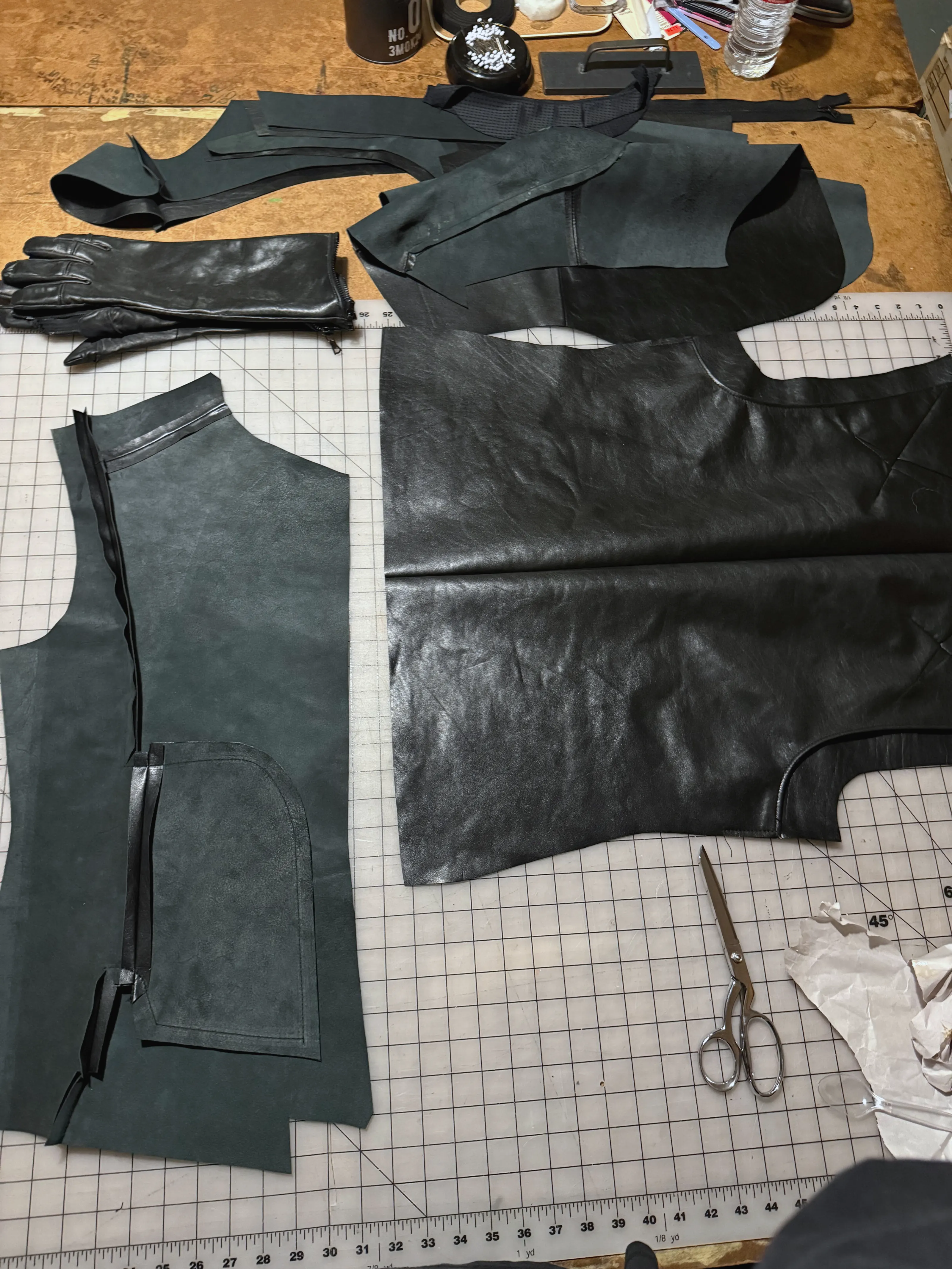 Black leather pattern pieces, a pocket-faced front panel and a long leather glove laid out on a cutting table with shears.