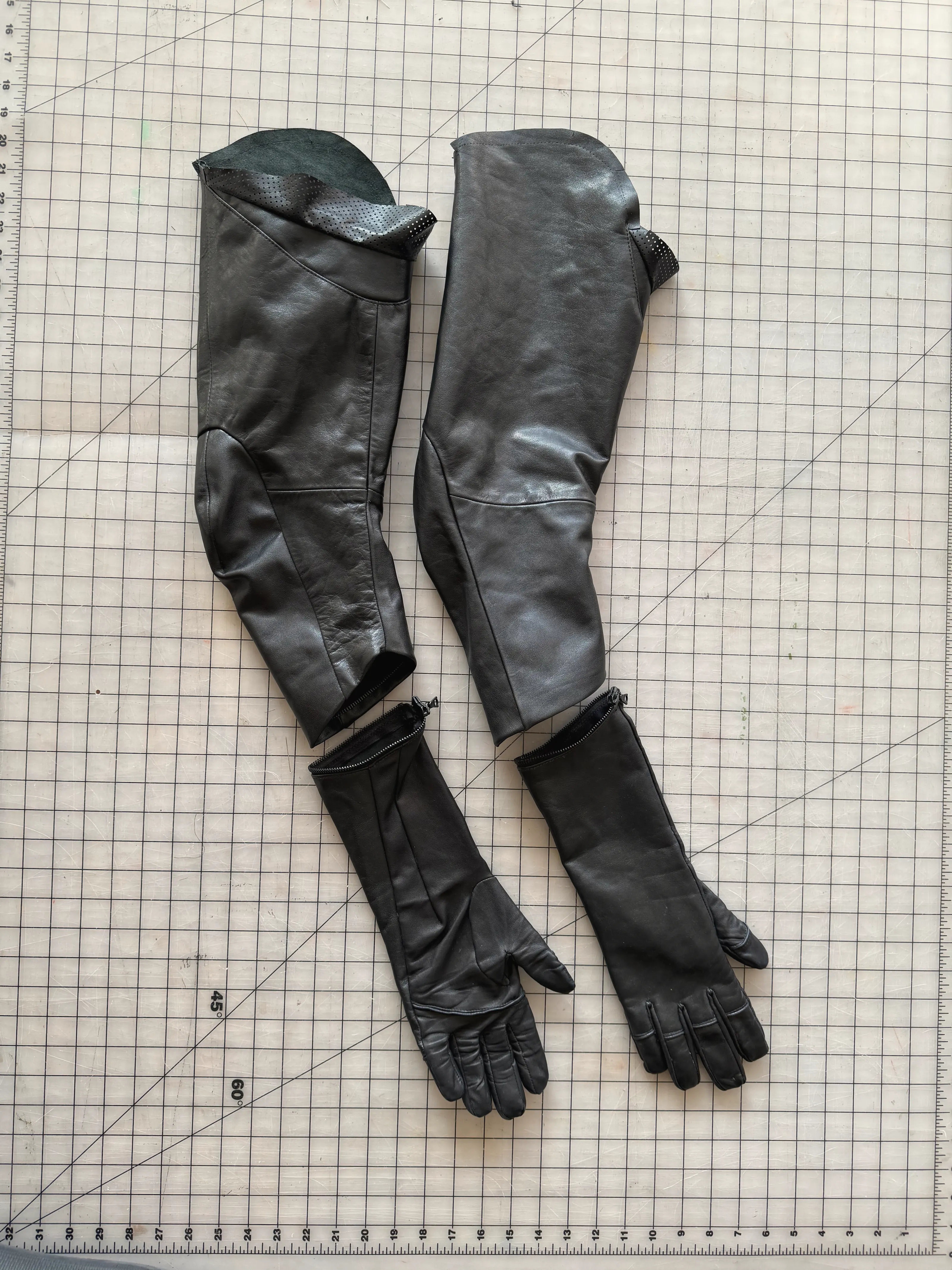 Pair of black leather sleeves with elbow seaming and perforated linings, each zipped at the cuff to a long leather glove, on a gridded cutting mat.
