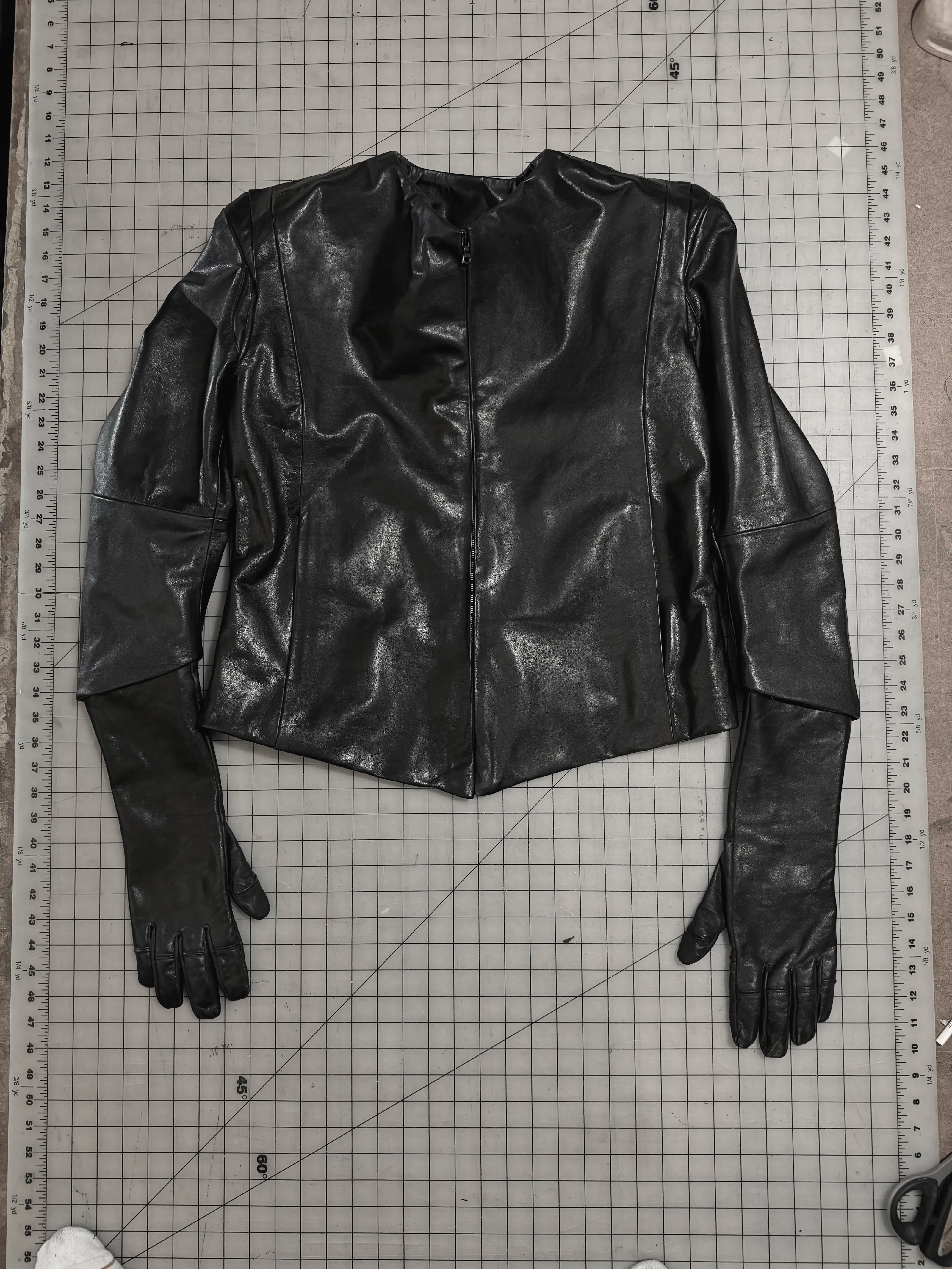 Cropped black leather jacket with a centre-front zip and long gloves integrated into the sleeves, laid flat with arms extended on a gridded cutting mat.
