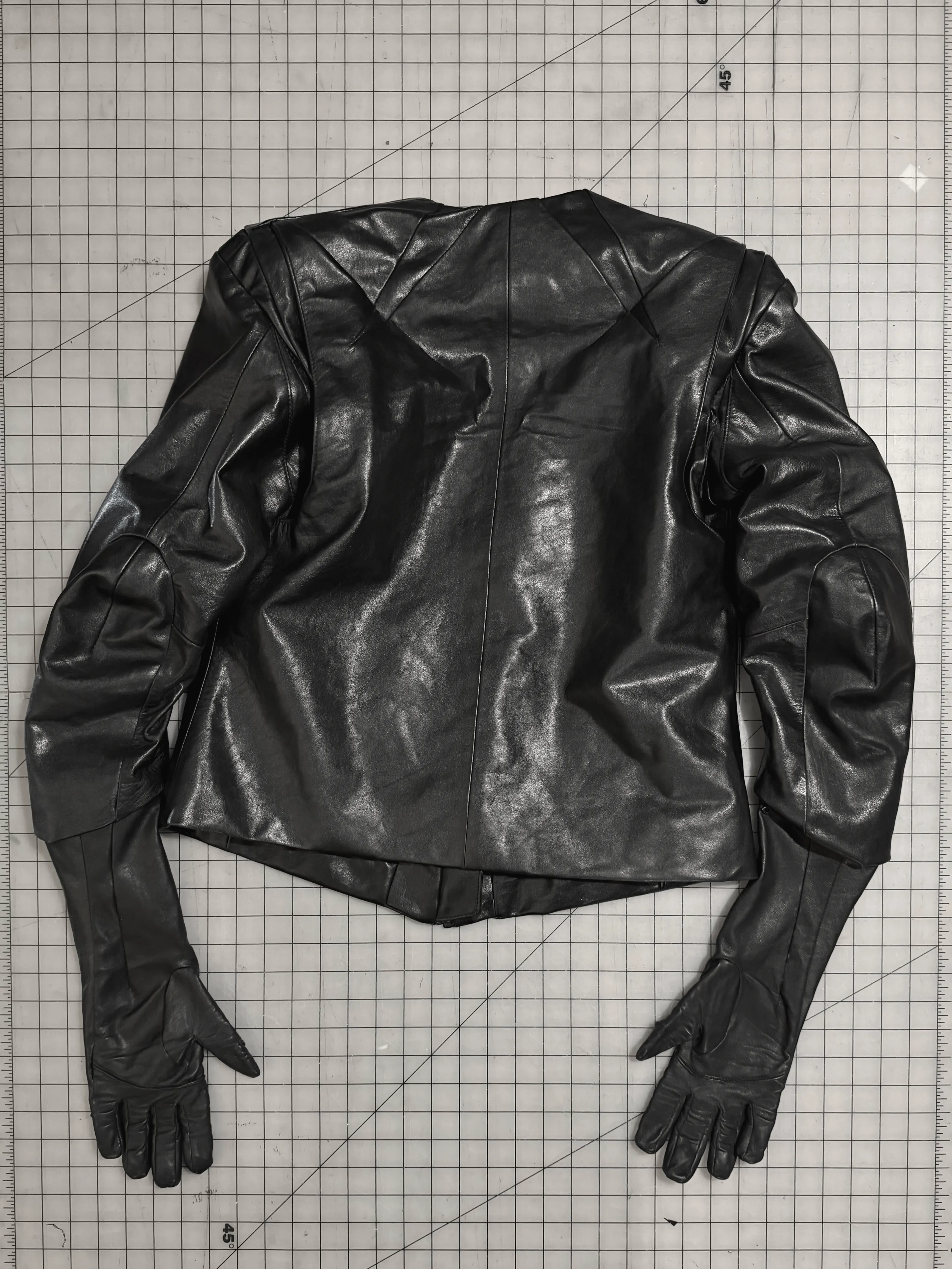 Cropped black leather jacket with integrated gloves, back view showing shoulder and elbow panel seaming, laid flat on a gridded cutting mat.