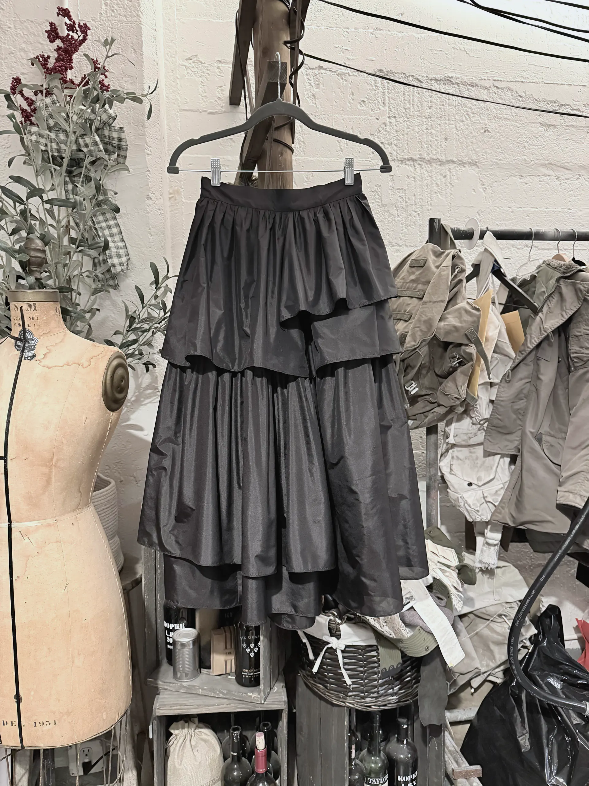 Black tiered taffeta skirt with a gathered waist, shown on a hanger in the studio beside a vintage dress form.