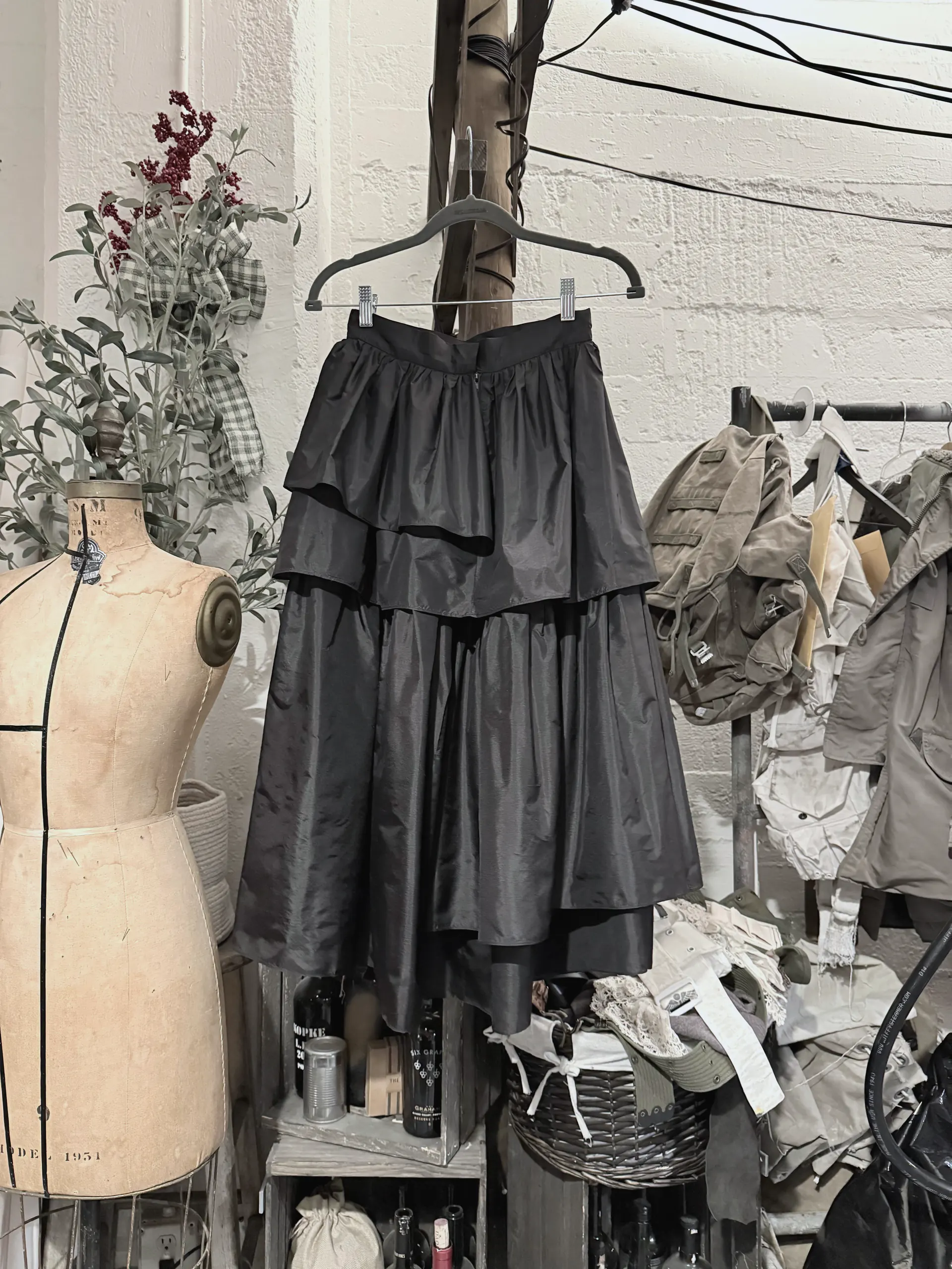 Black tiered taffeta skirt on a hanger, wider view showing the studio wall, dress form and rack of utility jackets behind.