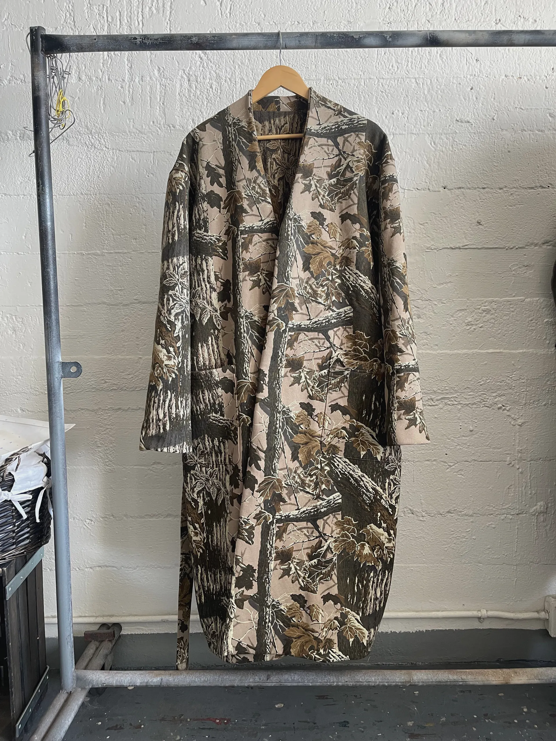 A full-length unstructured robe coat in tree-bark and leaf camouflage hangs open on a metal rail against a white painted brick wall. It has a collarless shawl-style front, long set-in sleeves and large patch pockets at the hip, and falls to the ankle.