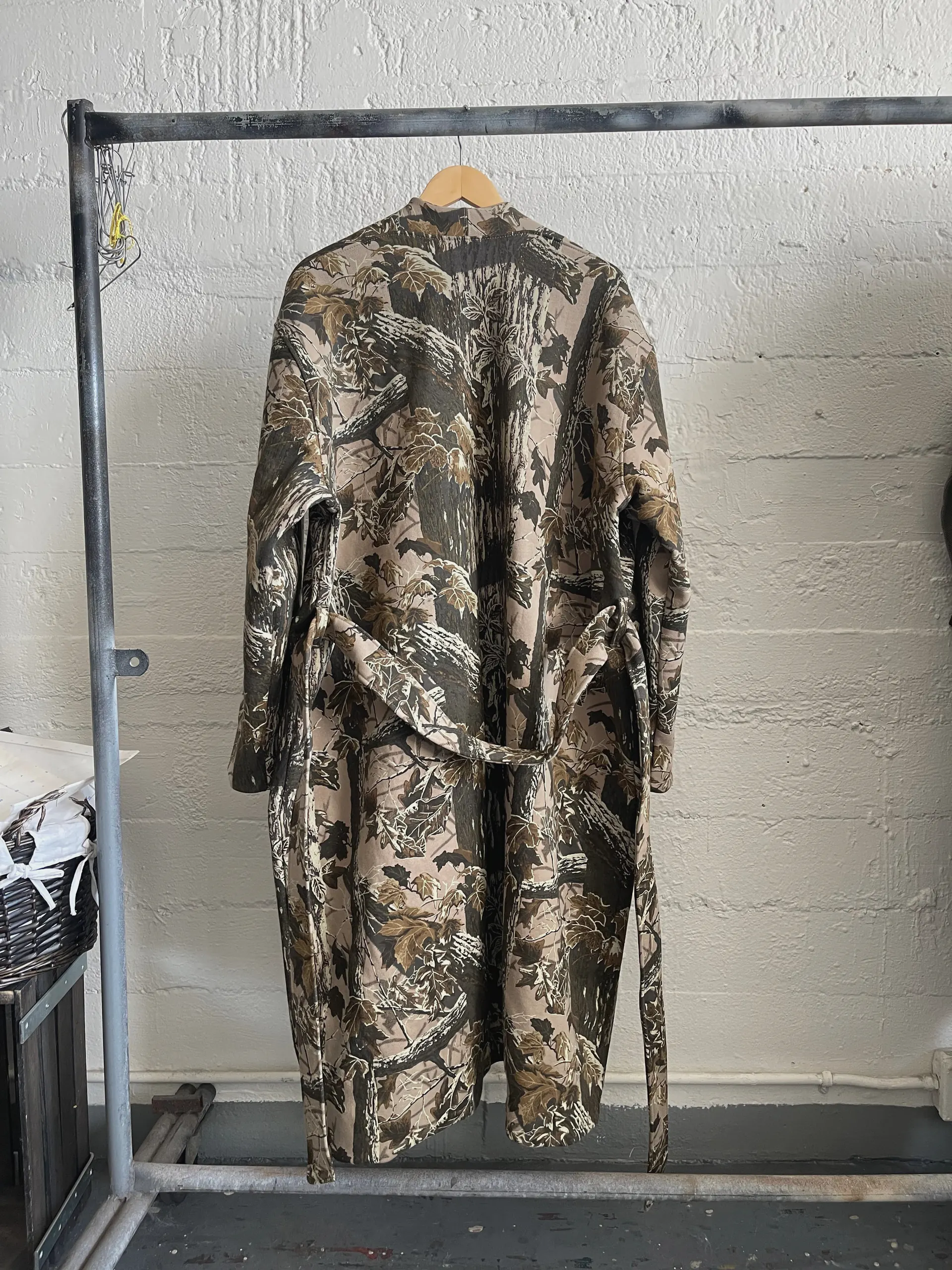 The camouflage robe coat is shown from the back on the same rail, the wide dropped shoulders and straight back panel falling to the ankle. A self-fabric tie belt passes through back loops at the waist and hangs loose down one side.
