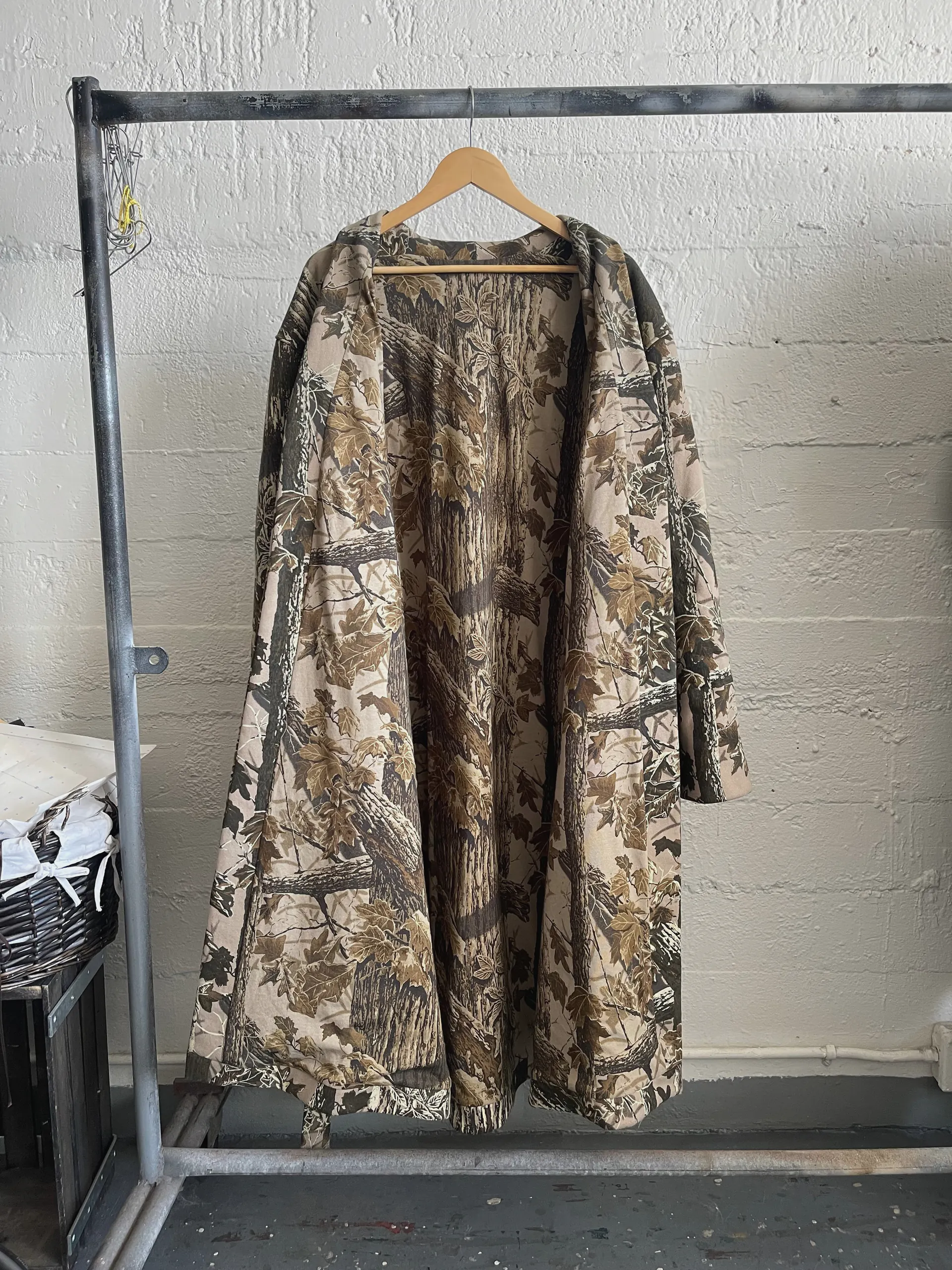 The camouflage robe coat hangs with both fronts swept back to reveal an interior faced in the same tree-bark print, so the coat reads as one continuous camouflage inside and out. The open drape shows the full sweep of the skirt and the depth of the hip pockets.
