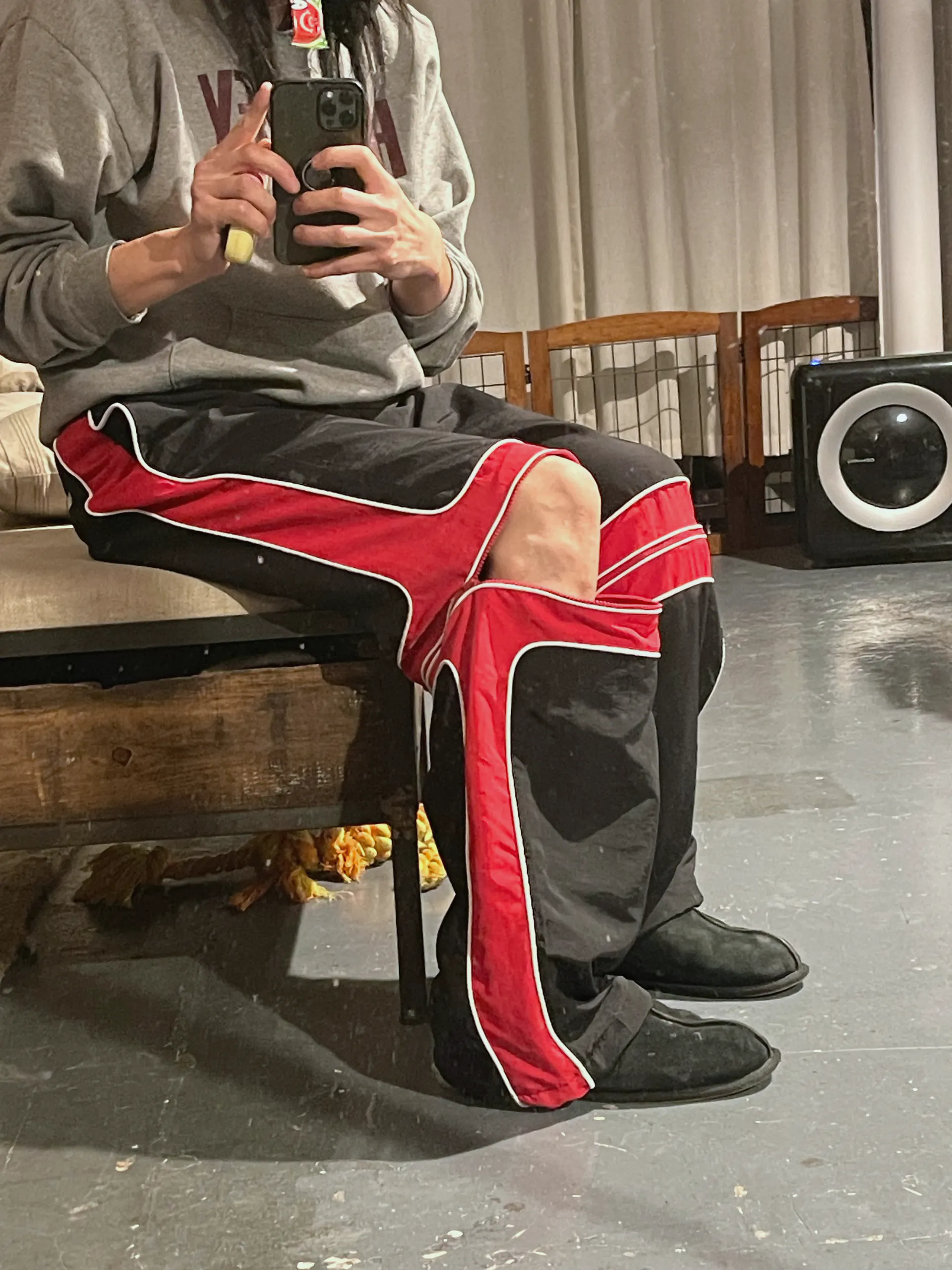 A mirror photograph of the black trousers worn seated, one leg crossed, with the red knee zip opened so the red band gapes and a bare knee shows through the split. The white-piped red panels run down the outside of both legs to the hem.