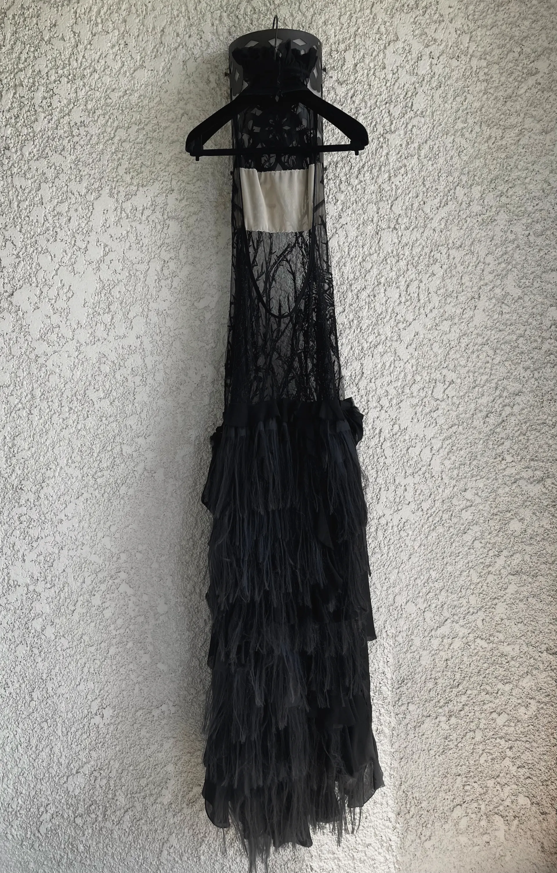 Full-length black gown on a hanger against a stucco wall: sheer embroidered lace bodice over an ivory panel, with a long tiered ruffled skirt.