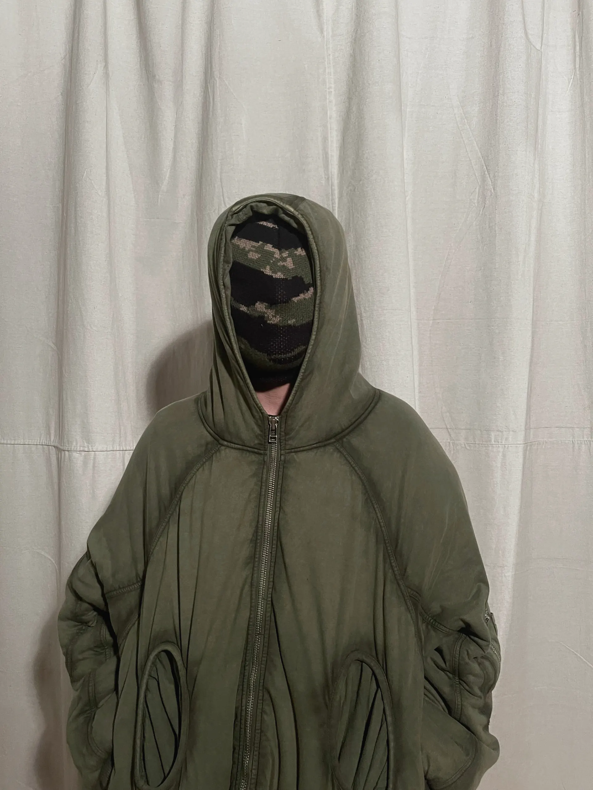 Oversized olive hooded jacket with a centre zip and sculpted oval pocket openings, worn front-on against a canvas backdrop.